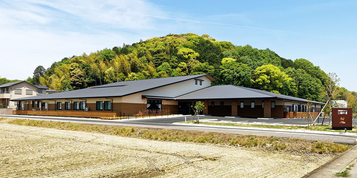 介護施設の外観。木造平屋建ての新築施設。背景に里山の緑。